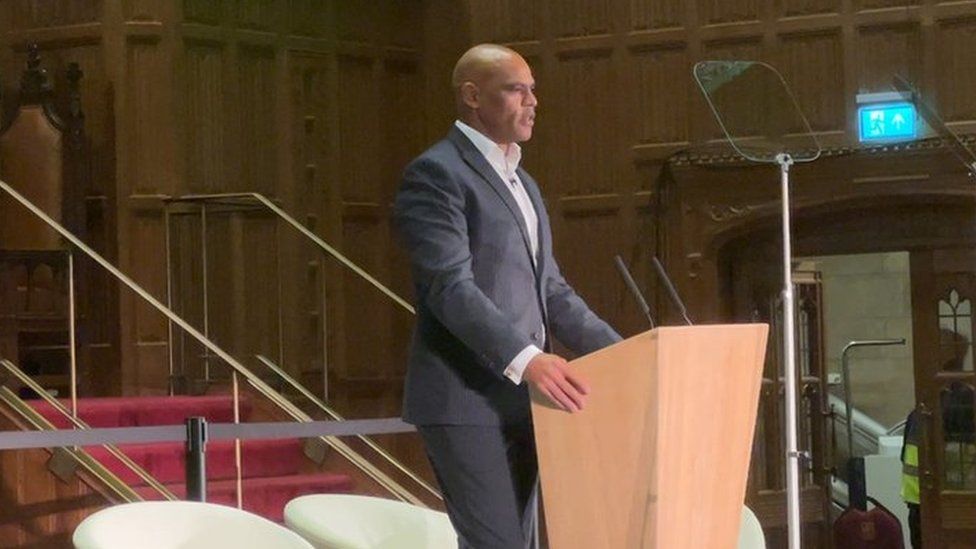 Marvin Rees with a bald head standing at a podium wearing a dark suit and a white shirt addressing a hall