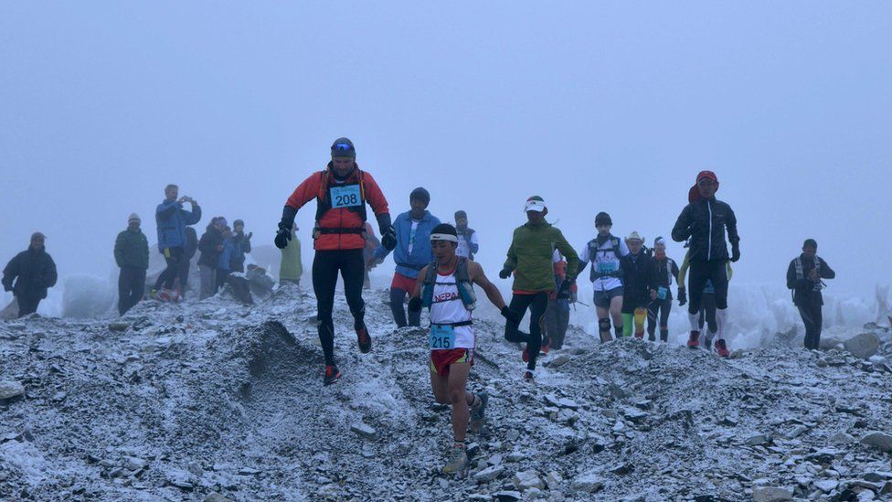 Runners dash over snowy, uneven terrain during the race, led by a Nepalese man in a vest and shorts, immediately followed by a foreign man in leggings and a warm coat and hat. 29 May 2016. Handout photo.