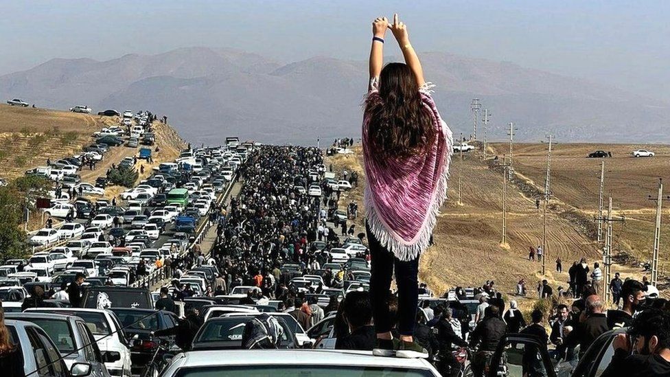 A young woman without a hijab stands on a car as a huge crowd walks towards the Aichi cemetery in Saqqez, Iran, to visit Mahsa Amini's grave on 26 October 2022