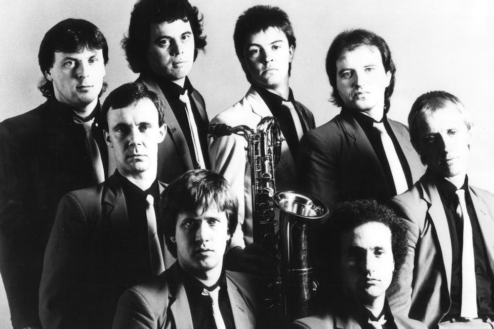 A black and white image showing members of Q Tips wearing dark shirts and light ties with a saxophone in the middle of the group.