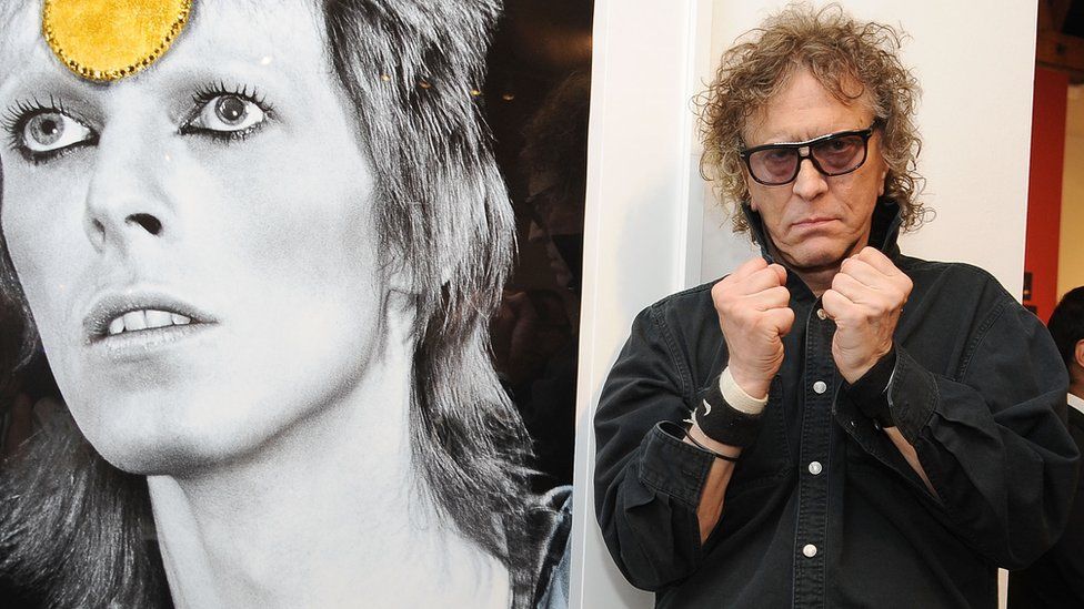Mick Rock in front of a picture of David Bowie