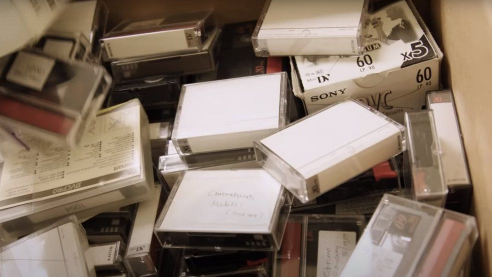 Крупный план коробки с различными небольшими видеокассетами. Они расположены хаотично — как будто их наскоро набросали друг на друга. Большинство из них в пластиковых футлярах для драгоценностей. Есть также картонные коробки меньшего размера, по-видимому, содержащие несколько лент с надписью «Sony, DVC, 60».