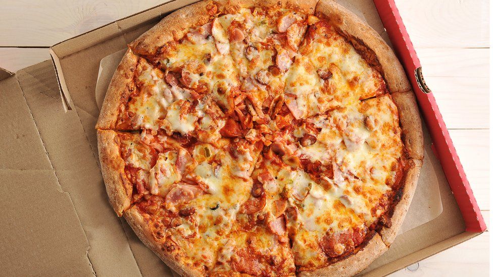 Overhead view of a take away pizza with cheese and spicy sausages in a cardboard box on a wooden table background.