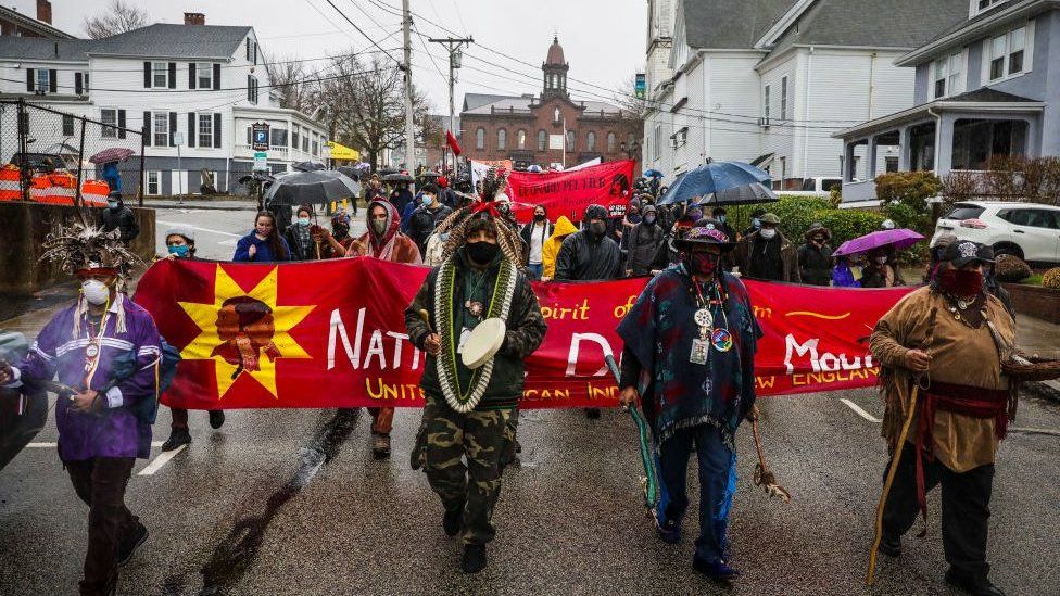 Demonstrators march through Plymouth on Thanksgiving in 2020, holding a large red banner that says 'National Day of Mourning.'