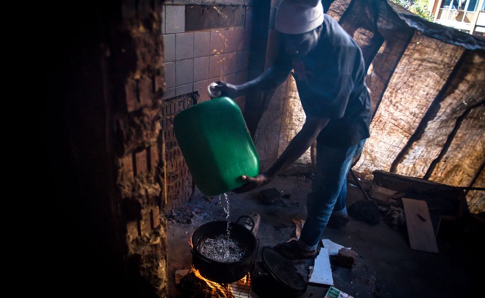 An unknown man pours water from a container into a pot over an open fire in preparation of cooking a meal in the derelict San Jose building in Johannesburg, South Africa