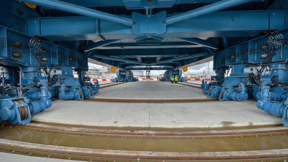 Construction workers stand underneath the underside and between four massive array of wheels on rails that maneuver "Big Carl",