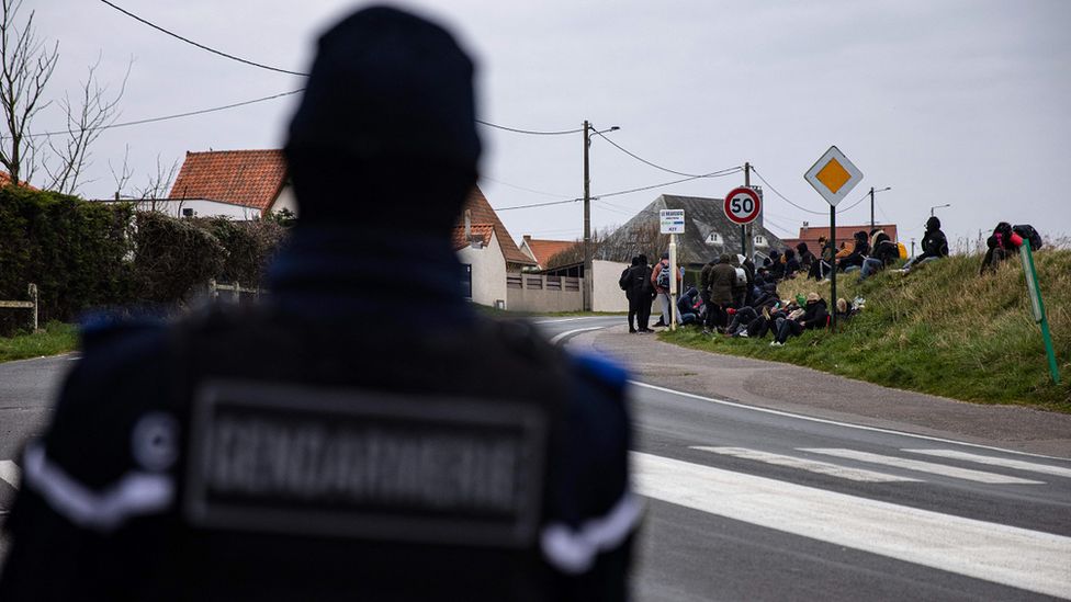 Французский жандарм стоит рядом с группой мигрантов, ожидающих автобус в Кале после неудачной попытки пересечения границы в марте 2022 года