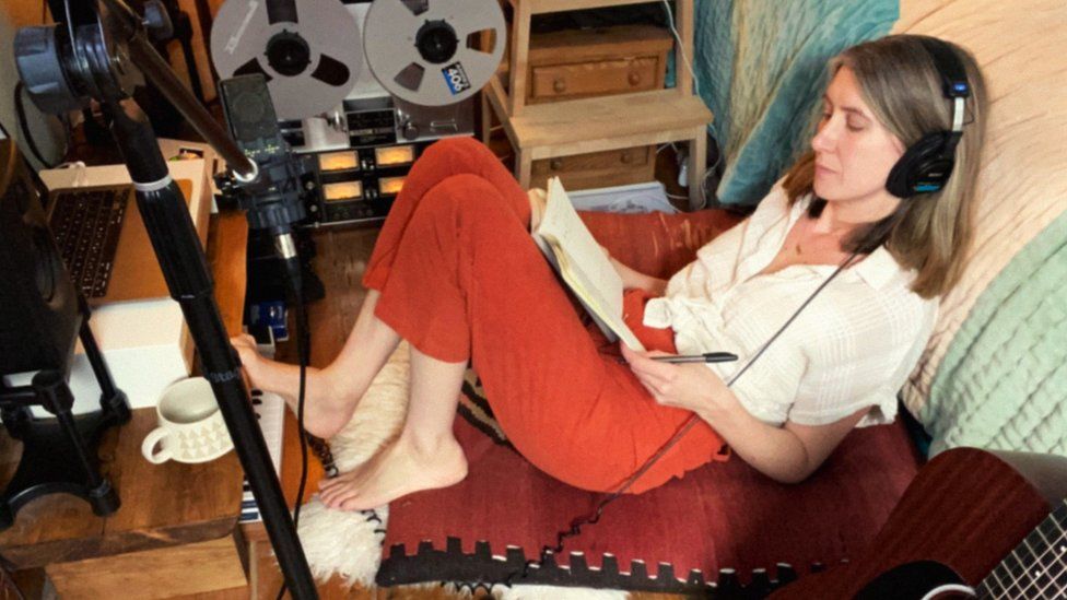 image of Jenny Lindfors. She is pictured sat on the floor, wearing headphones and writing in a notebook. A microphone stand and guitar can be seen in the shot.