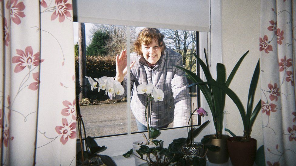 Mavis' friend Heather waves through the window at her