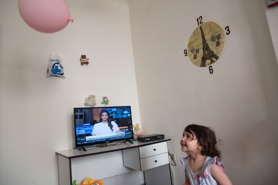Fatima, 4, is playing with a balloon at her new home in Chania. Fatima is Ahmed's youngest daughter. On the background, the cable TV is showing the news from a Syrian TV channel.