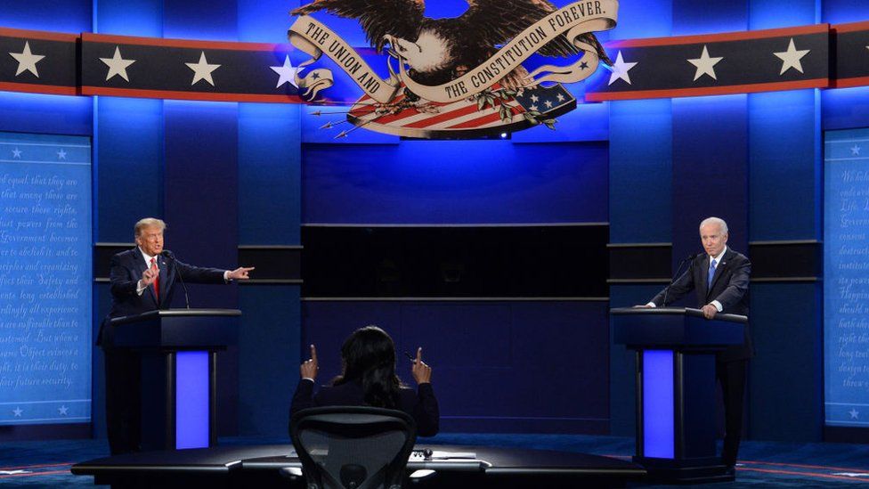 Donald Trump and Joe Biden standing behind podiums on stage during a debate while a woman with her back to the camera moderates