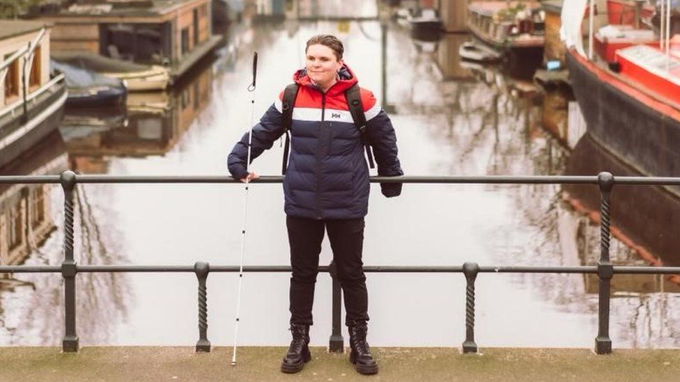 Connor Scott-Gardner stood wearing black boots and black trousers with a navy, white and red coat and his white cane leaning against a bridge over a canal