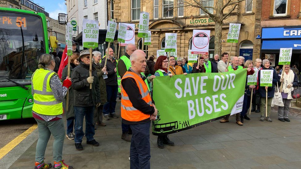 A group of people holding a large green banner reading: "Save our buses"