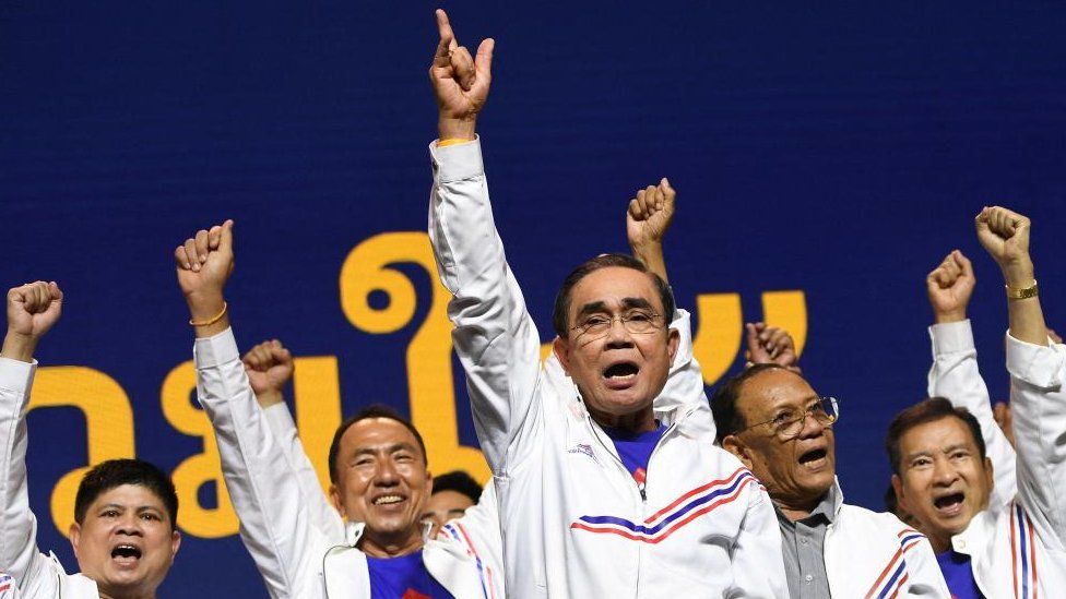 Led Prayuth Chan-ocha, a group of men in white jackets raise their arms