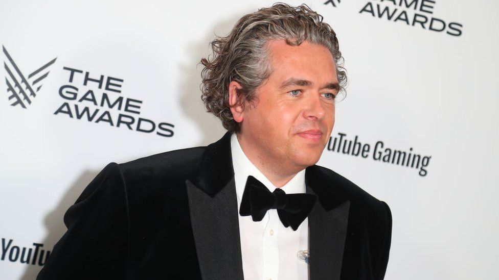 A man with medium-length grey hair, brushed back, wears a tuxedo with black jacket, white shirt and black bow tie as he poses on a red carpet. He's in front of a white hoarding with logos for The Game Awards and YouTube Gaming printed across it in a repeating pattern. He casts a faint shadow on the board as a result of light from the photographers' flash bulbs hitting him