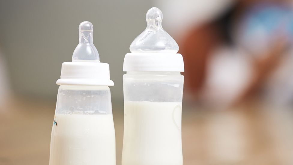 Two bottles for feeding babies milk in focus - a mother and baby, out of focus, behind - stock shot
