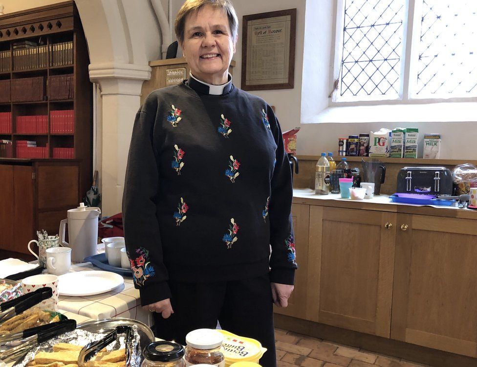 A female vicar standing inside a church with tea cups and cakes on a table
