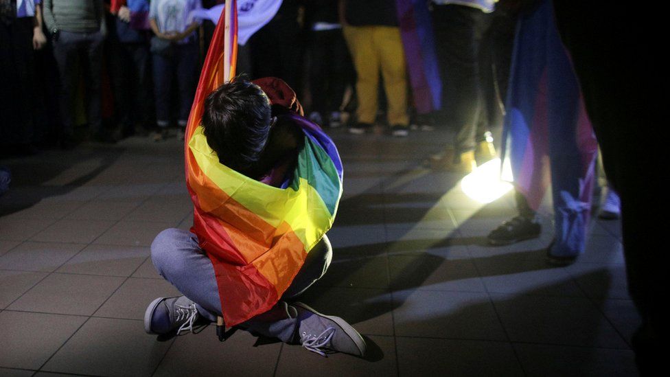 A man wrapped in a rainbow flag sits alone on a tiled floor, head bowed