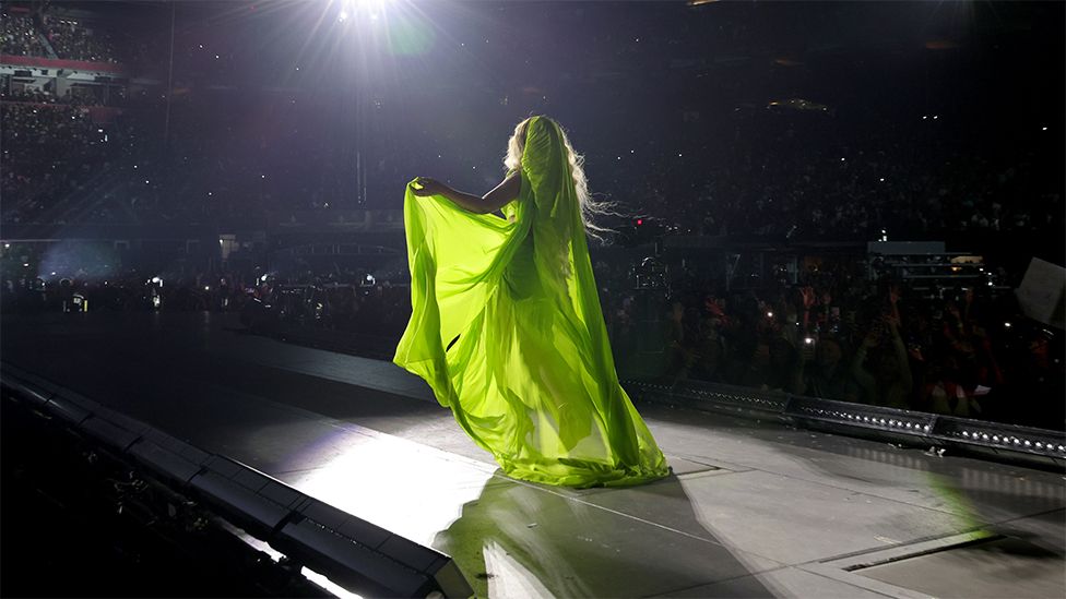 Beyoncé' wearing Gaurav's design. The dress is neon green and has a flowing hood and a knotted design on one shoulder. Pictured from the back, the sheer dress is floor length with a train and the singer holds some material in her left hand.