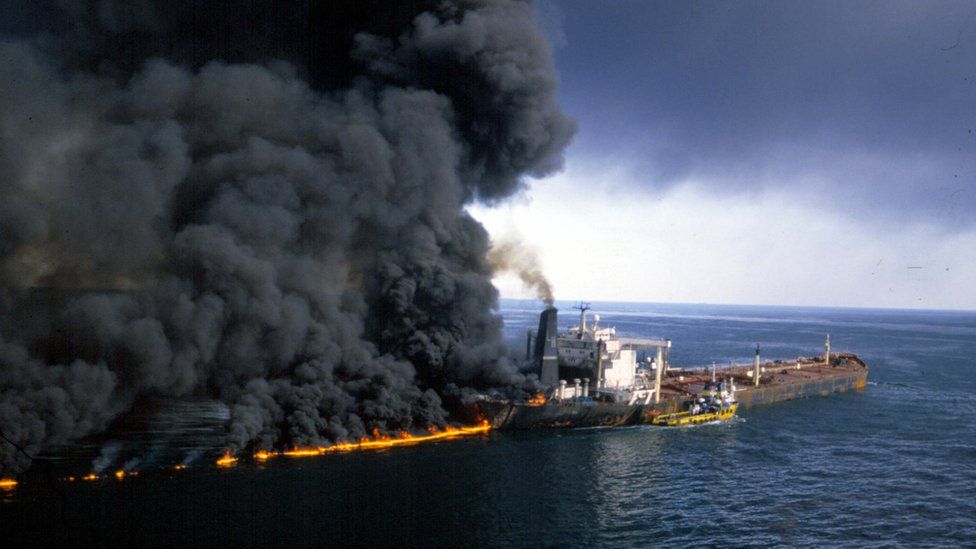 On an open sea, a large industrial ship is seen burning, with thick clouds of black smoke billowing out of it