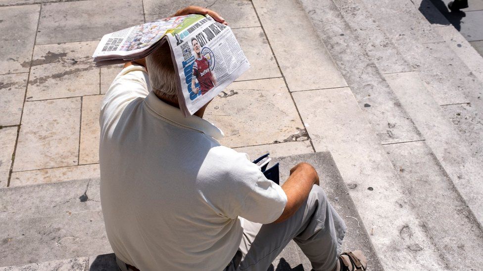 A man holds newspaper over his head to shade himself