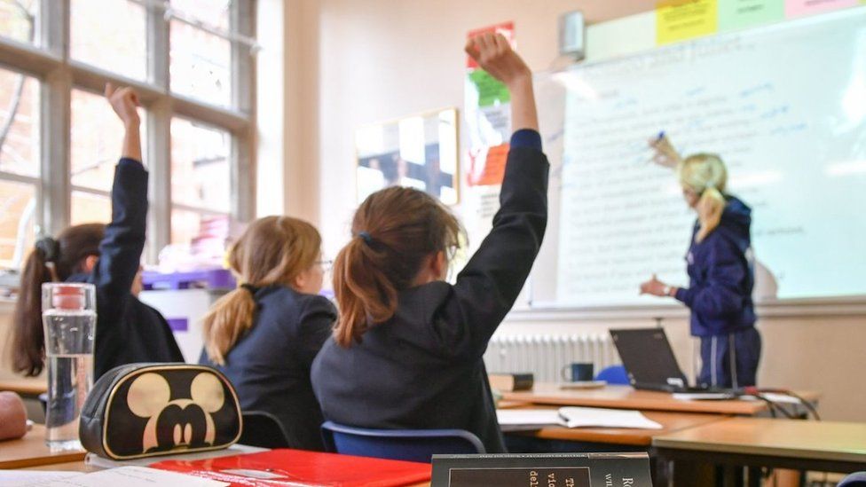 File image of girls in a classroom holding their hands up in front of a teacher and whiteboard
