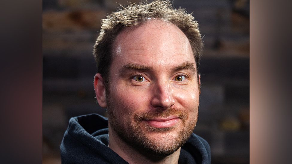 A man with shortish, wispy brown hair wears a blue hooded top in a headshot-type picture. He has shortish brown hair, the strands illuminated by the camera's flash. He also has a short stubbly beard. He's smiling slightly. Behind him is a brown wall suggesting a warehouse or trendy office space.