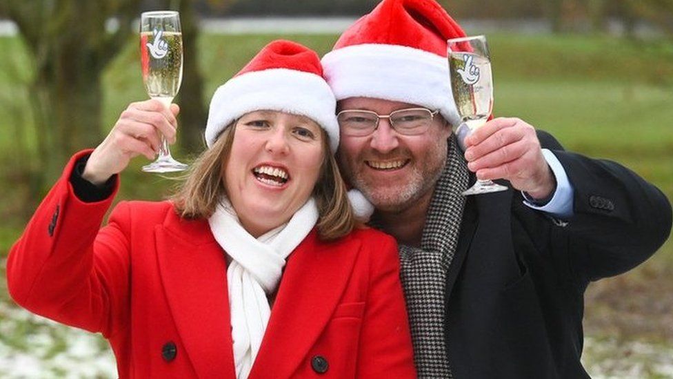 Ellie Land and Karl Ward, wearing Christmas hats, celebrating their lottery win with glasses of champagne