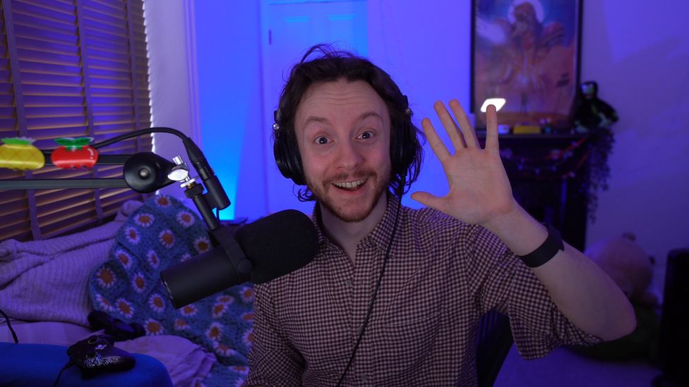 A man smiles, waving at the camera. He's in a room set up for livestreaming, equipped with a microphone on a boom arm and with lighting that lends it a blue hue.