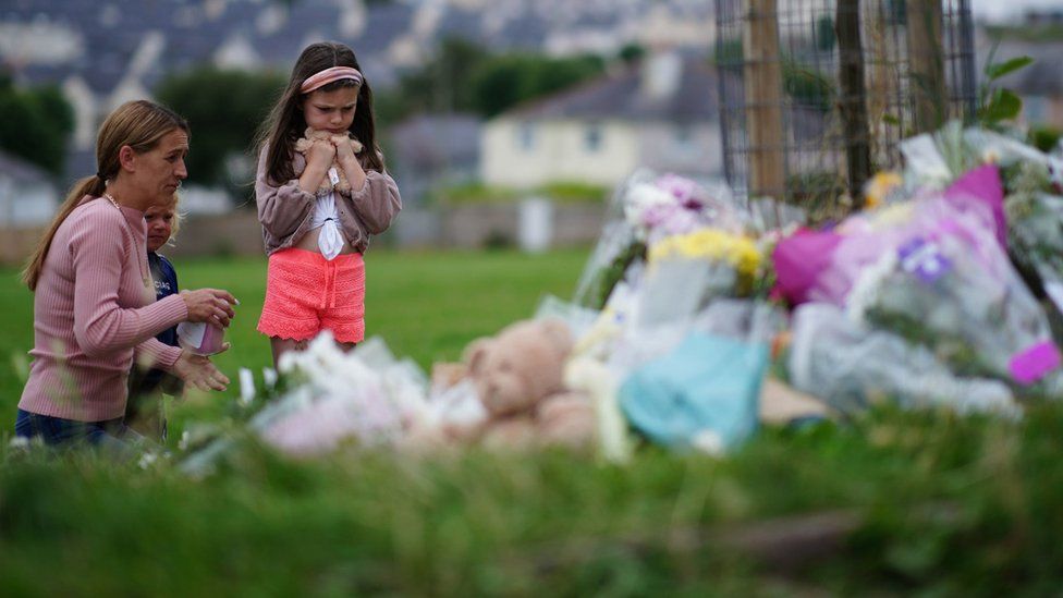 A woman and two children look at floral tributes to the five victims