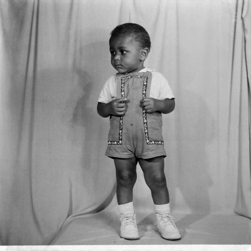 A toddler wearing a T-shirt, dungarees and lace-up shoes poses inside the camera studio.