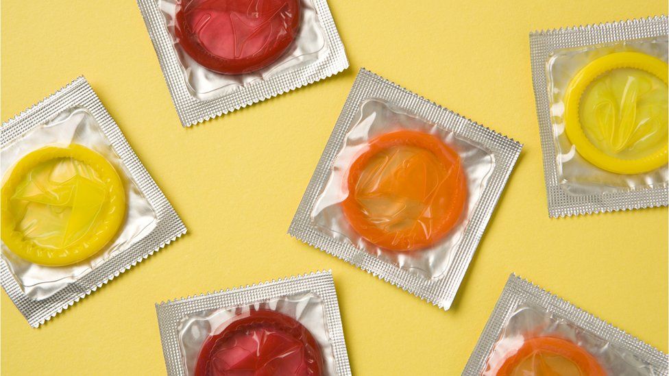 Stock image of condoms - six coloured condoms that are yellow, red and orange wrapped in silver foil are placed against a pale yellow background