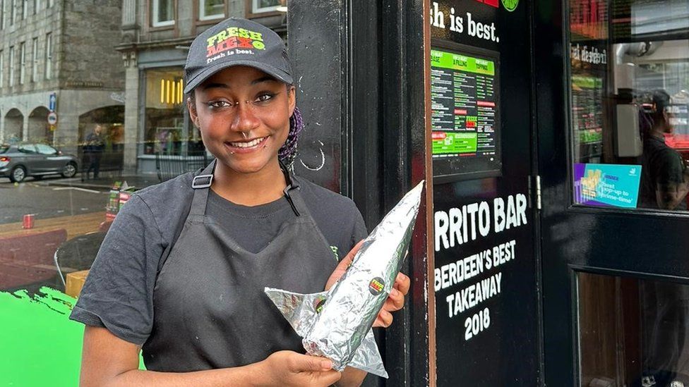 Anastatia pictured at her work at a burrito bar in Aberdeen. Ana is a young black woman with her hair worn in long braids, some coloured white and purple, tied back in a pony tail and worn beneath a work cap. She also wears a grey work apron over a grey T-shirt. She has piercings on the bridge of her nose, her septum and her lip and is smiling at the camera. She holds a burrito wrapped in foil to resemble a rocket ship.