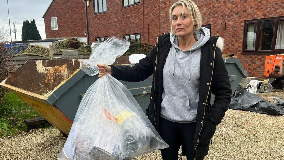 Kate Smith from the MHRA holds up a bag of evidence. She's a white woman with short blonde hair and wears a black parka over a grey hoodie. She's pictured outside a property with a skip outside