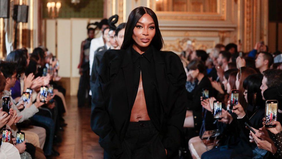 Naomi Campbell walking in Torishéju's show at Paris Fashion Week in October 2023. Naomi is a 51-year-old black woman with her long hair perfectly straight. She wears a black shirt with exaggerated shoulders unbuttoned except for at the collar, exposing her midriff. She has her hands in the pockets of the black trousers it's paired with. The show is in a very grand room with wooden flooring and gold detailing. The runway is between audience members, many of whom are filming the collection on their smartphones