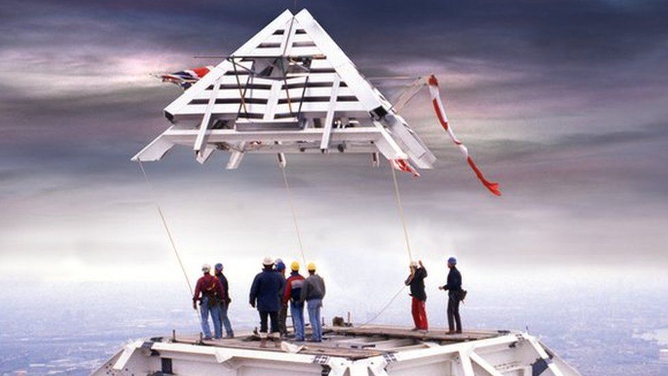 The pyramid-shaped top of One Canada Square, decorated with flags, being lifted into place by crane while workers look on.