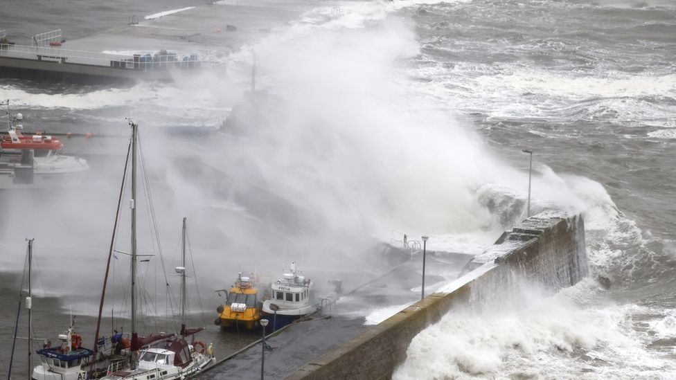 Photograph of large waves overtopping a harbour area crashing over moored boats.