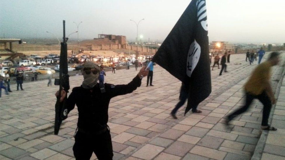 A fighter of the Islamic State of Iraq and the Levant (ISIL) holds an ISIL flag and a weapon on a street in the city of Mosul, 23 June 2014.