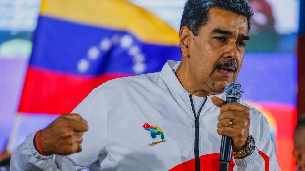 Venezuela's President Nicolas Maduro addresses supporters at a rally. He is wearing a white tracksuit top. The country's flag is seen over his shoulder.