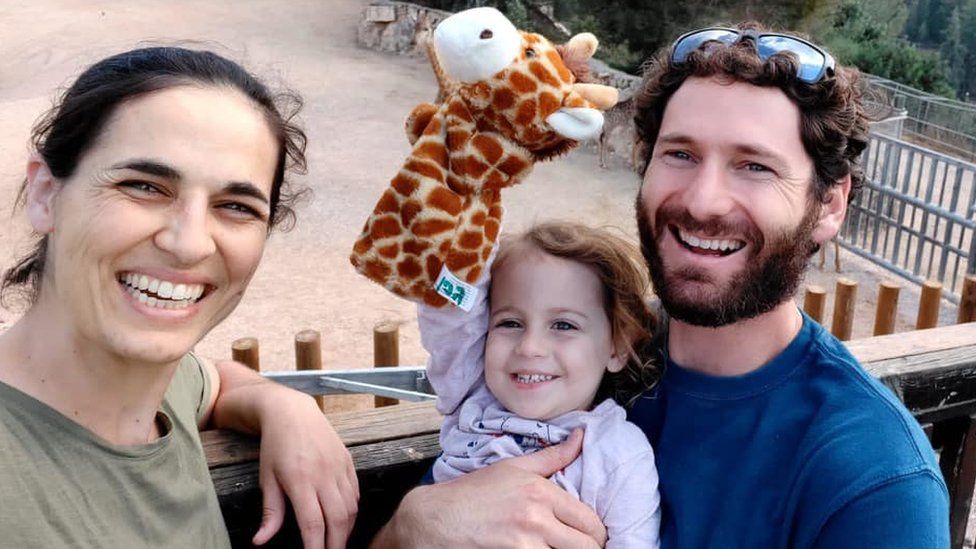 A photo of Yarden Roman-Gat, her husband Alon and three-year-old daughter Gefen, smiling as the child holds up a giraffe glove puppet