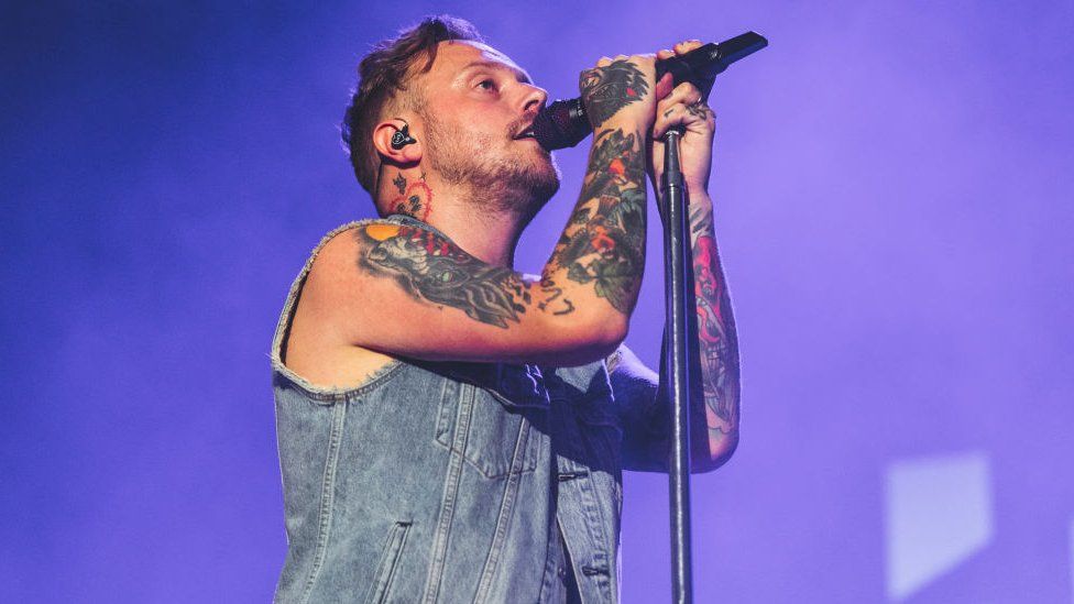 Sam Carter, lead singer of Architects. Sam is a white man in his 30s with short brown hair and stubble. He has a heart-shaped tattoo on his neck and heavily tattooed arms which he holds up around a microphone stand. He wears a sleeveless denim jacket and and is pictured singing on stage which is lit purple.