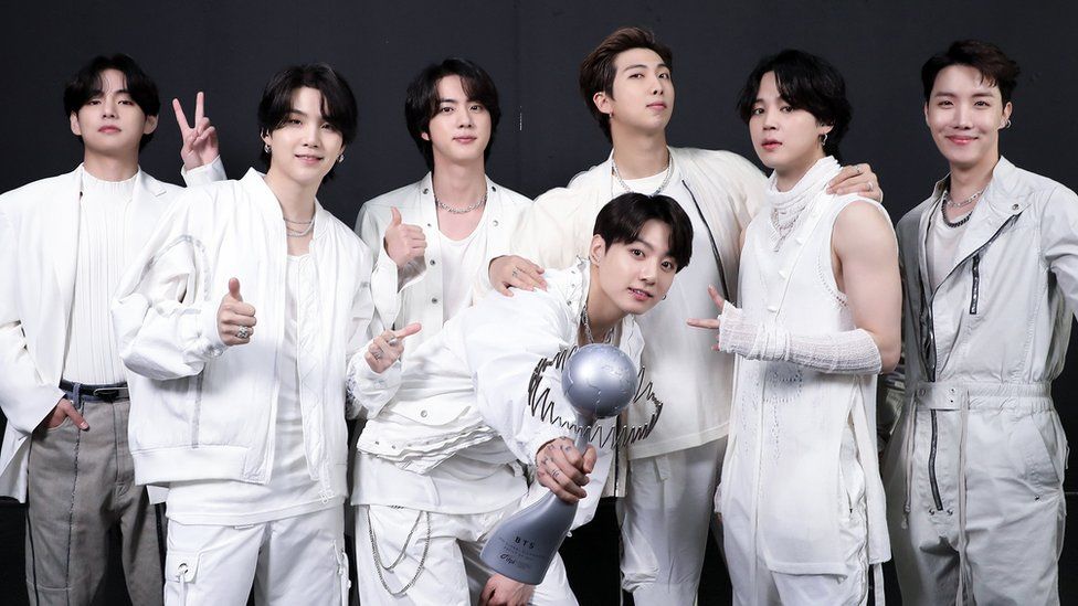 Все семь участников BTS позируют для фото, получив награду. Корейский бойз-бенд, одетый в белое, позирует с улыбающимися лицами, на черном фоне позади всех семерых