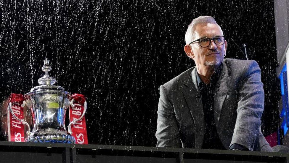 Gary Lineker in a rainy stadium by the FA Cup trophy