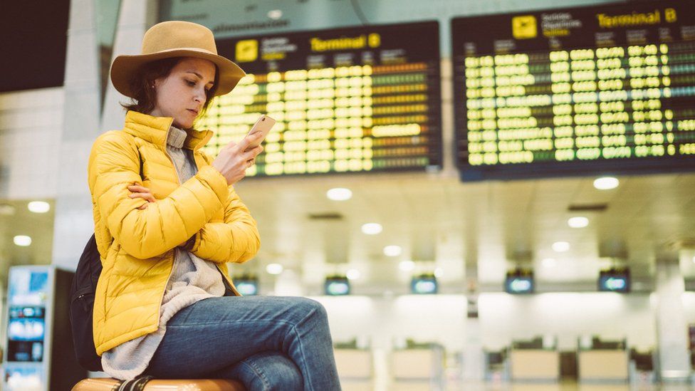 A woman sits on her luggage and looks at her phone in front of an airport departures board