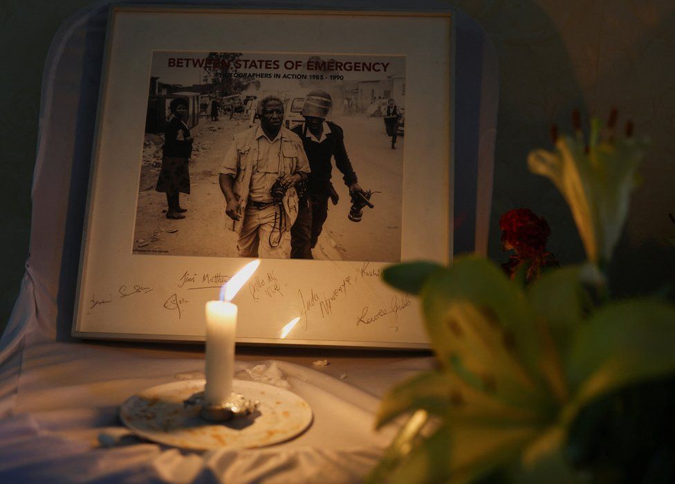 A candle burns next to a framed photograph showing the late Peter Magubane being arrested decades ago.