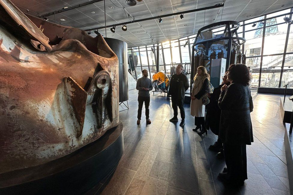 Five people stand next to a large buckled piece of metal in a museum