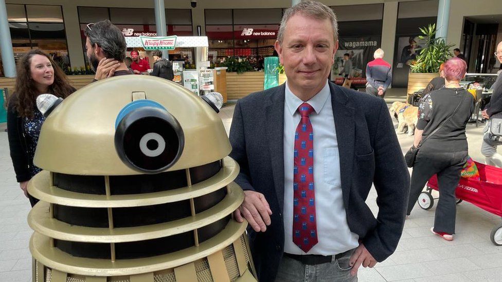 A man stands next to a gold dalek in a busy shopping centre.