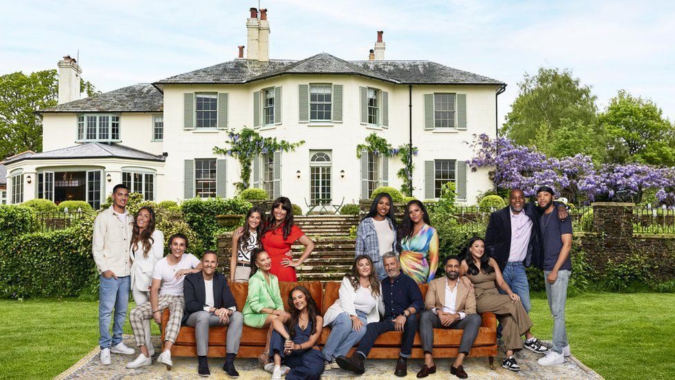 The cast of the ITV dating show My Mum, Your Dad pose in front of the country house where the show is set. They are grouped in their pairs so children with the mum or dad. Four of the pairs are sat on the brown sofa which is on a large rug on a green grass lawn. The other four pairs are stood behind the sofa. Lots are leaning on the other person or have their arm around then. Behind them are some stone steps and bushes, and then a grand white country house.