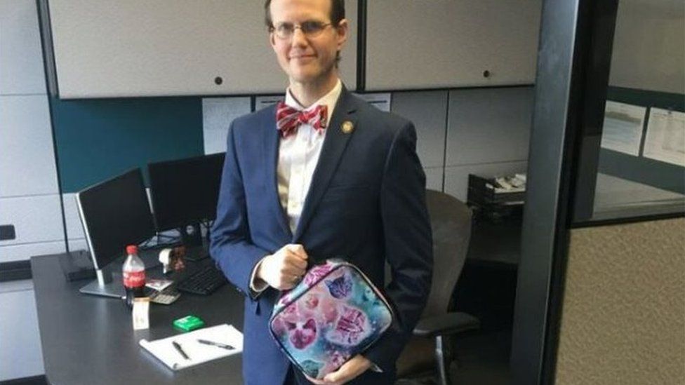 photo shows David Pendragon wearing a suit in an office and holding a lunchbox with pictures of cats on it.
