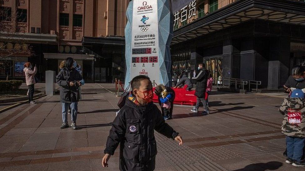 Parents with their children wearing face masks stand next to a countdown display showing days left for the Beijing 2022 Winter Olympics, on a shopping street in Beijing, China, on 17 January 2022